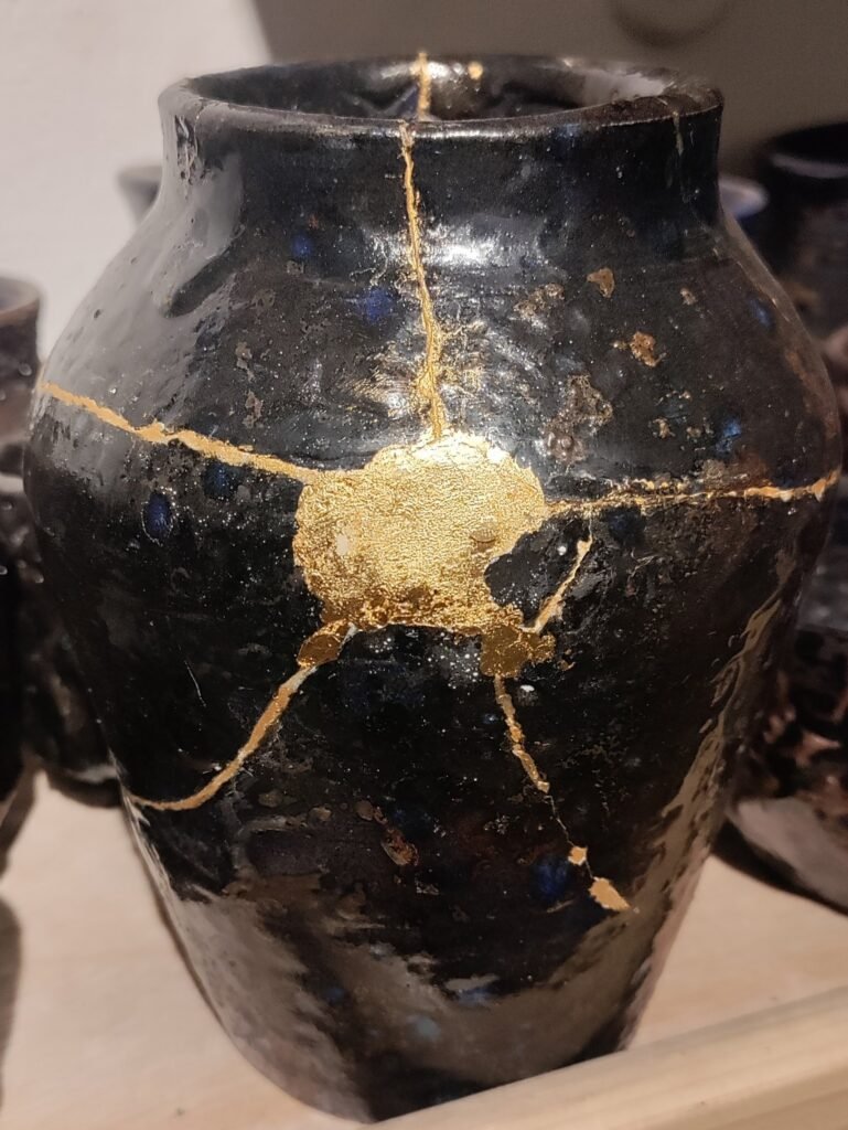 A dark ceramic jar repaired with kintsugi showing gold seams and a gold filled area where a chip was lost